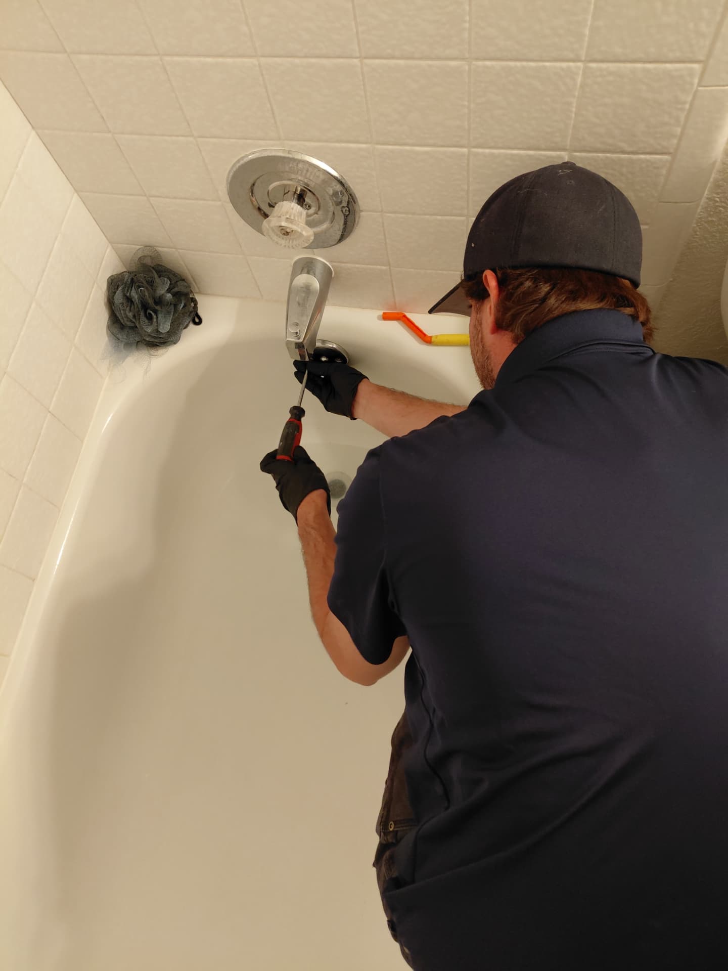 A BluDog technician in black gloves using a screwdriver on the chrome overflow plate of a white bathtub in a tiled bathroom.