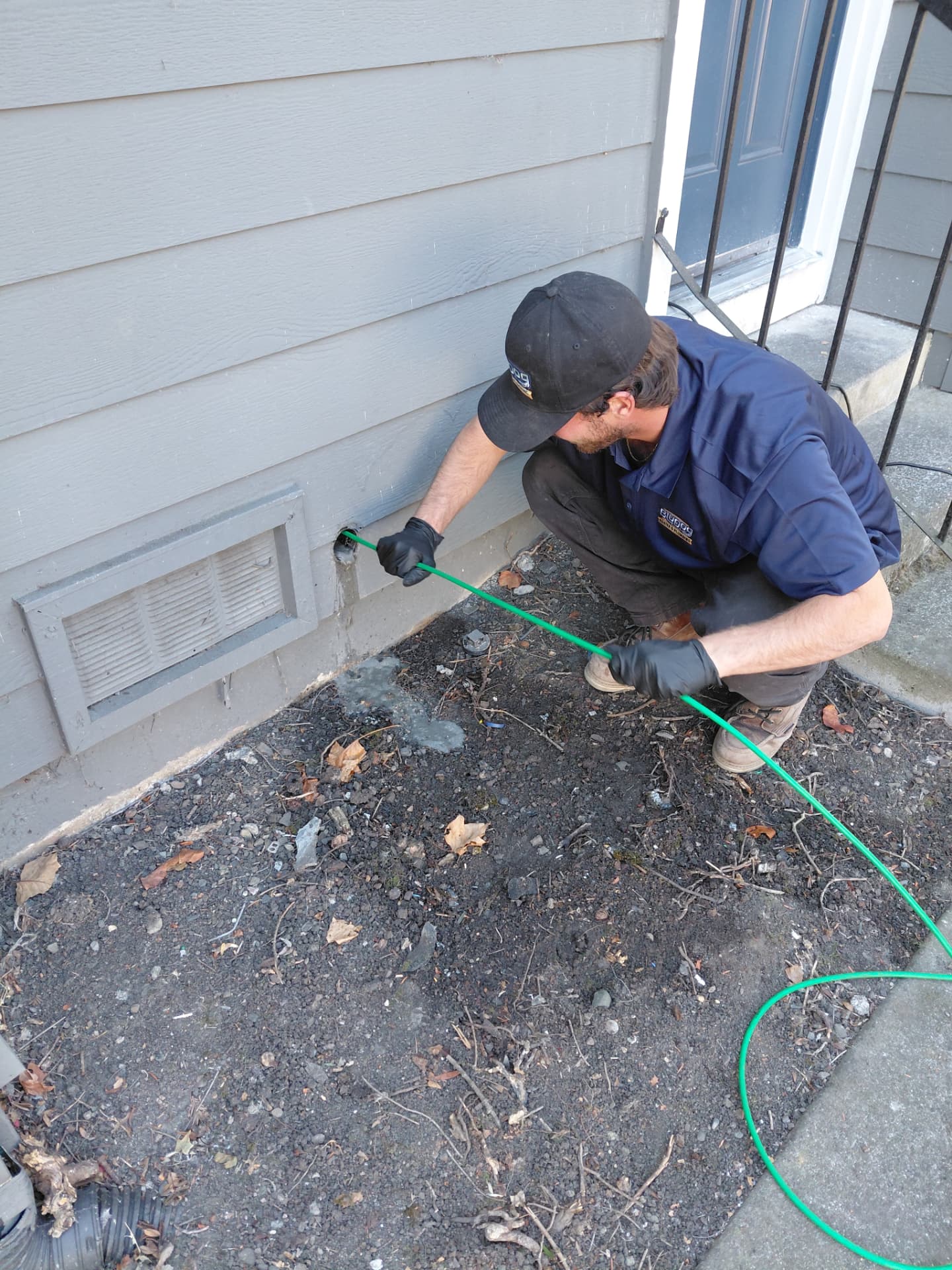 A BluDog technician crouched beside a home’s foundation, feeding a green drain-clearing line into an exterior cleanout pipe.